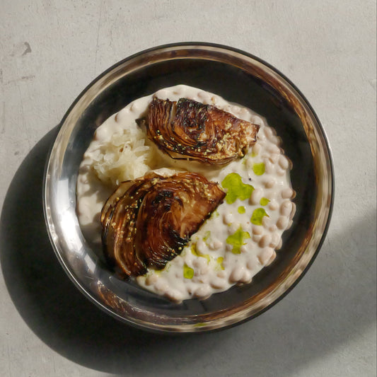 Two cabbage wedges on a bed of creamy sauce with green oil in a silver bowl on a light gray background